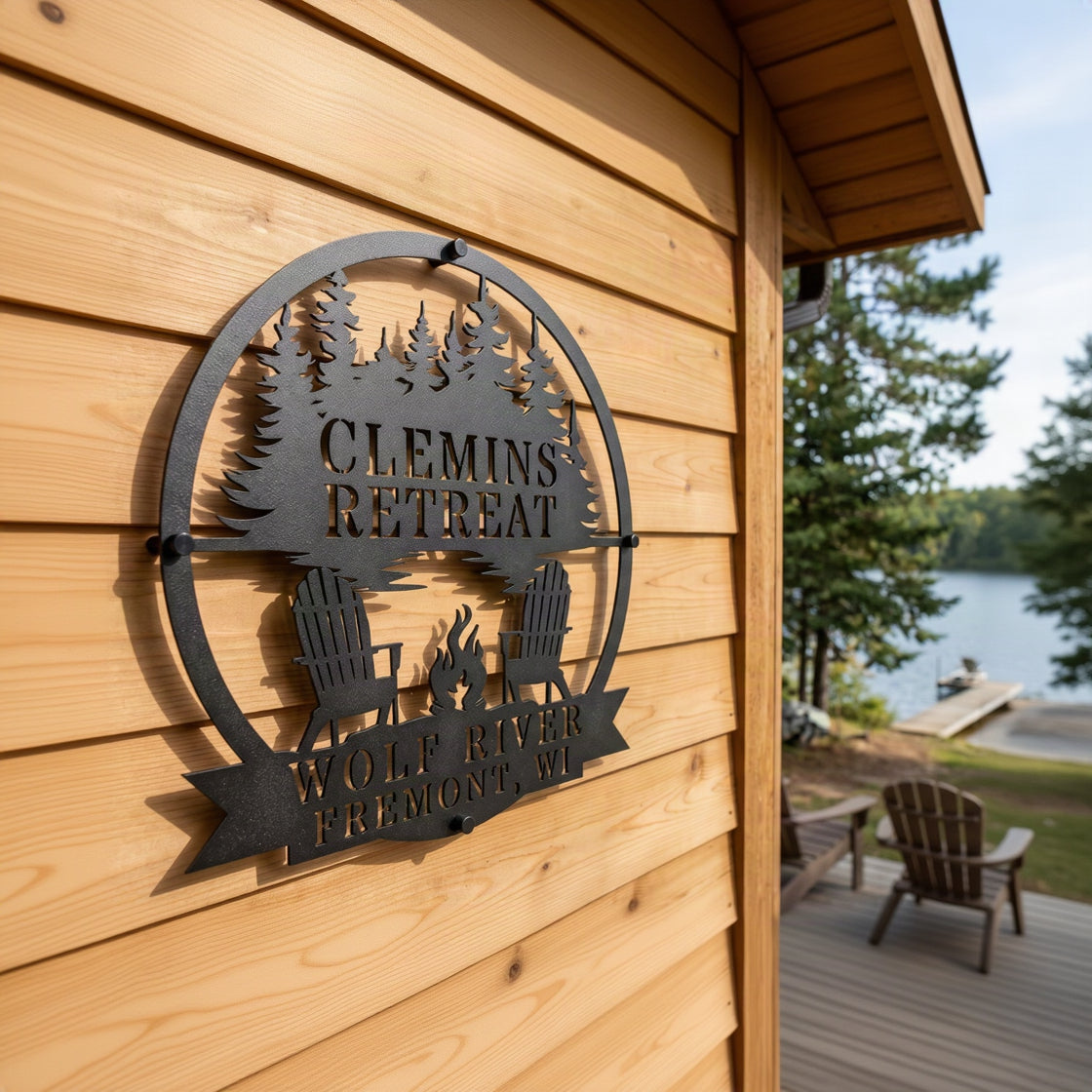 Customized campfire by the lake metal wall art sign in matte black, mounted on light cedar wood cabin siding with a scenic lake view in the background.