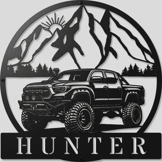 Custom offroad Toyota truck metal sign in powder-coated matte black, featuring mountain peak backdrop and personalized name plate, mounted on a light gray studio wall.